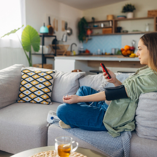 Junge Frau sitzt auf dem Sofa und misst ihren Blutdruck w&auml;hrend sie auf ihr Smartphone schaut.