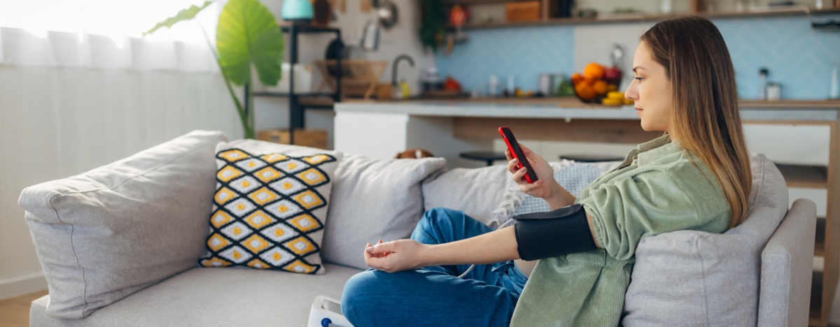Junge Frau sitzt auf dem Sofa und misst ihren Blutdruck w&auml;hrend sie auf ihr Smartphone schaut.