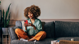 Eine gl&uuml;ckliche Frau, die auf ihrem Handy schreibt, w&auml;hrend sie auf dem Sofa sitzt