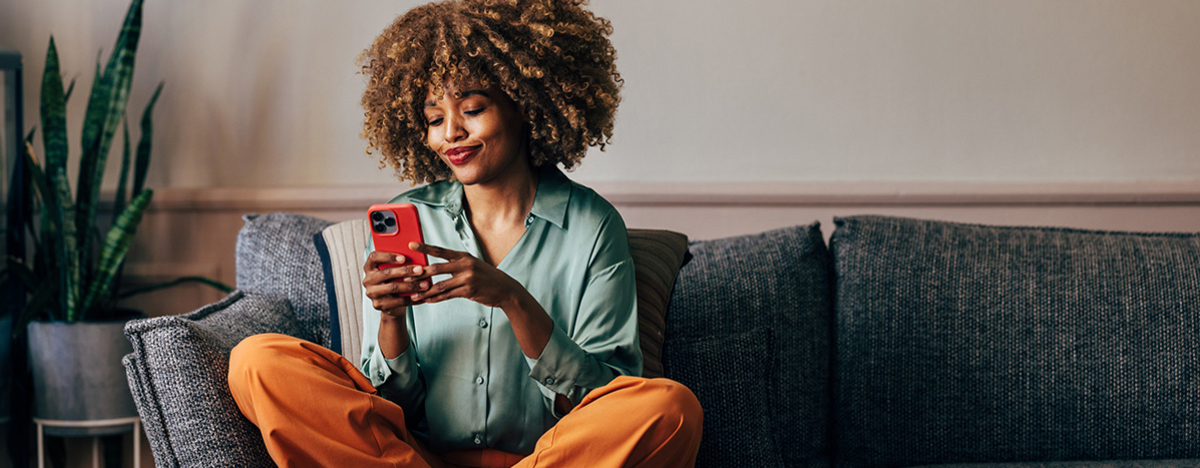 Eine gl&uuml;ckliche Frau, die auf ihrem Handy schreibt, w&auml;hrend sie auf dem Sofa sitzt
