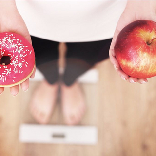 Frau steht auf Personenwaage und h&auml;lt in der einen Hand einen Apfel, in der anderen einen Donut