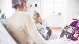 Eine pflegebed&uuml;rftige Frau sitzt im Bett, eine Pflegerin zieht ihr spezielle Schuhe an