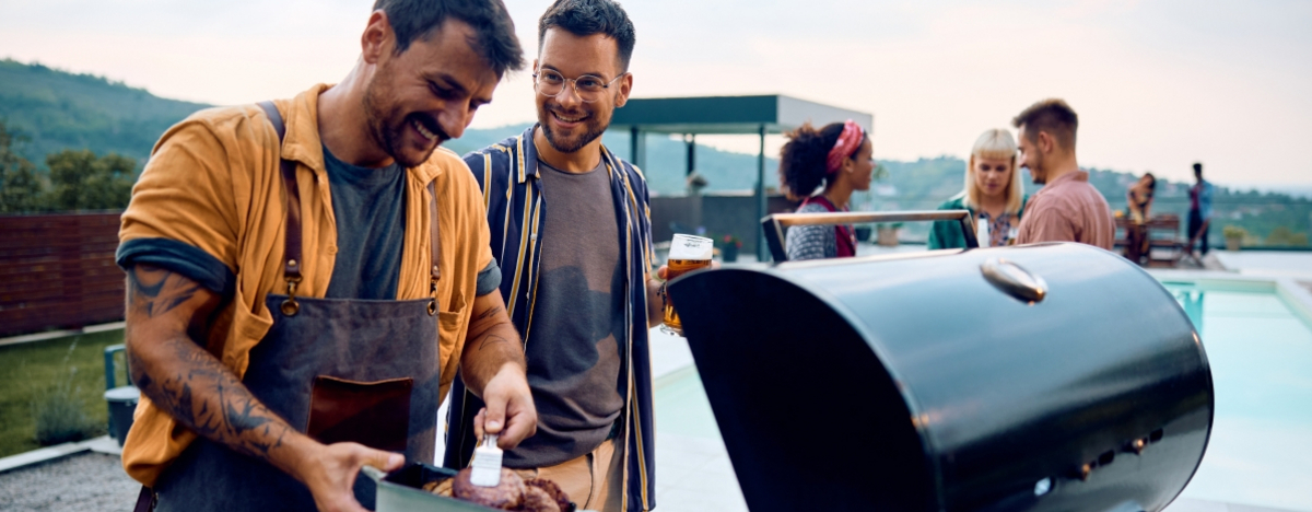 Zwei M&auml;nner stehen am Grill und lachen