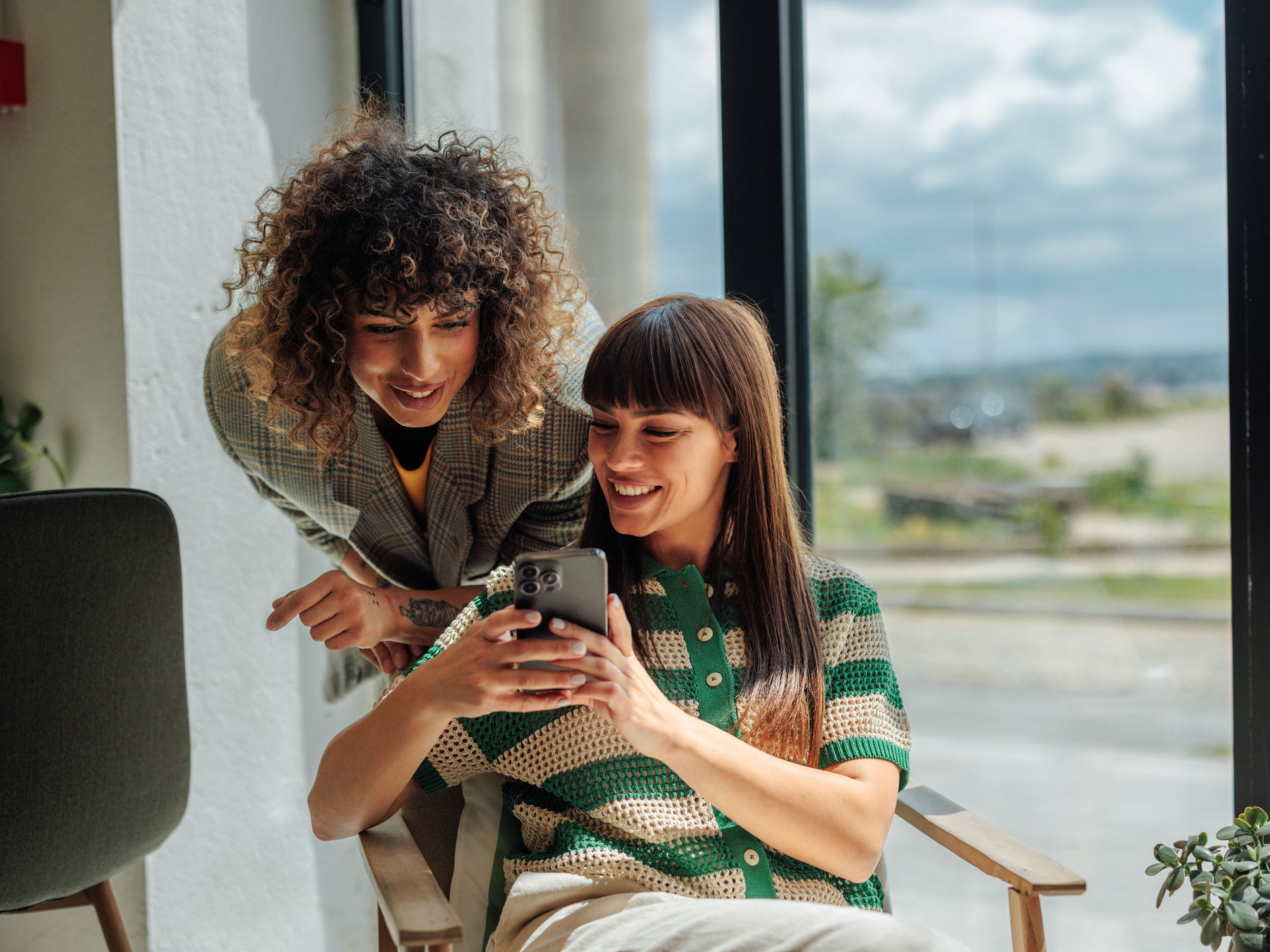 Zwei Frauen sitzen vor einem gro&szlig;en Fenster und schauen interessiert auf ein Smartphone.