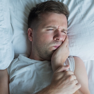 Mann liegt mit Zahnschmerzen im Bett und h&auml;lt sich eine Hand an die Wange