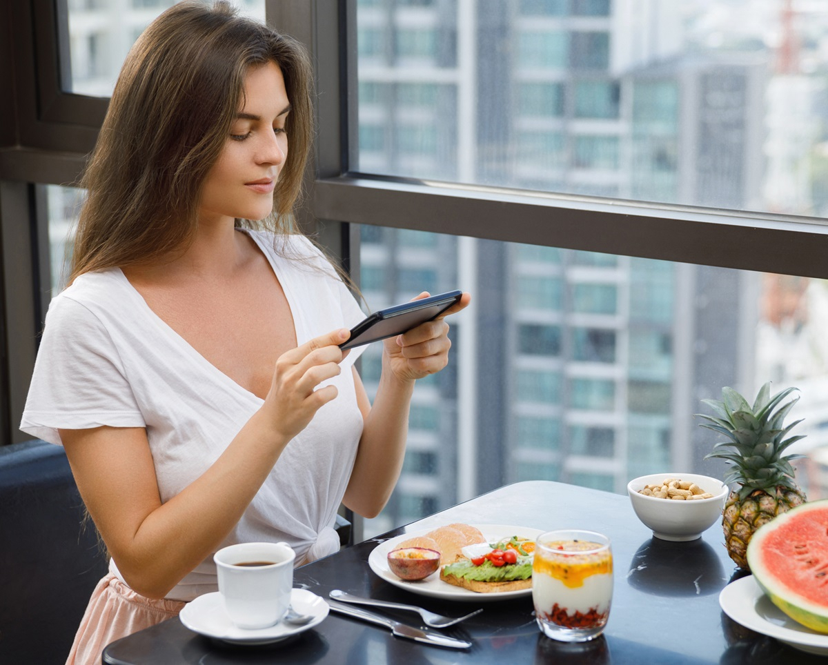 Das leckere Fr&uuml;hst&uuml;ck mit Joghurt, Obst, Kaffee und Toast wird von junger Frau f&uuml;r Social Media fotografiert