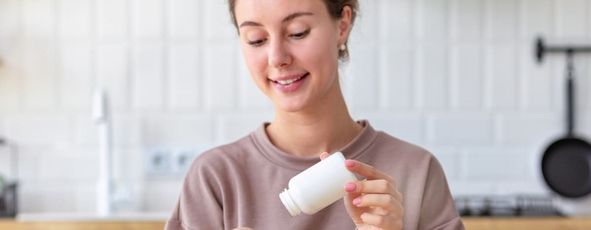 Vitamine, Nahrungserg&auml;nzungsmittel, gesundes Lebensstilkonzept. Junge Frau, die fr&uuml;hst&uuml;ckt und Vitamin zu sich nimmt, sitzt zu Hause in der K&uuml;che und l&auml;chelt freundlich