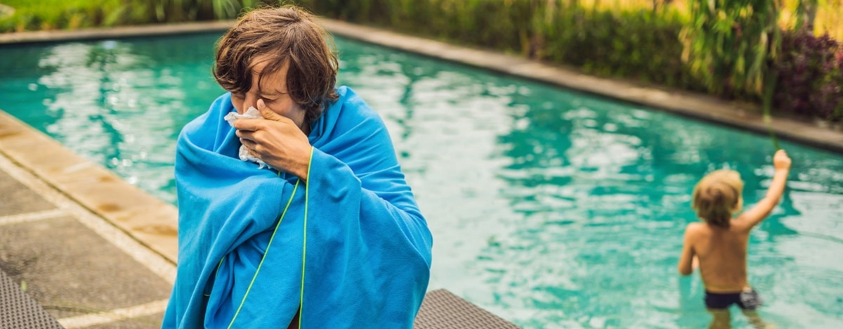Kranker Reisender. Der Mann hat sich im Urlaub erk&auml;ltet, sitzt traurig am Pool, trinkt Tee und putzt sich die Nase in eine Serviette. Sein Sohn ist gesund und schwimmt im Pool.