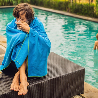 Kranker Reisender. Der Mann hat sich im Urlaub erk&auml;ltet, sitzt traurig am Pool, trinkt Tee und putzt sich die Nase in eine Serviette. Sein Sohn ist gesund und schwimmt im Pool.