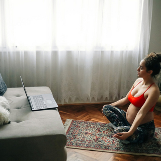 Schwangere Frau beim Yoga zu Hause und vor ihr ein Laptop
