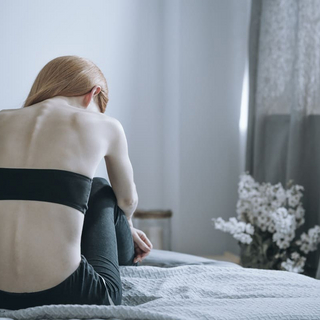 Eine junge Frau mit Magersucht sitzt auf einem Bett, mit dem R&uuml;cken zur Kamera.