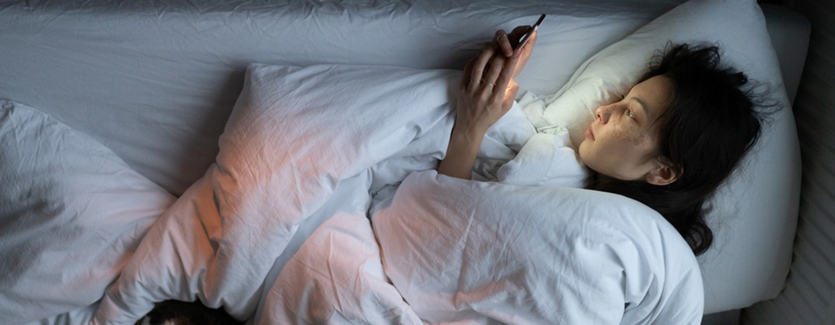 Eine junge Frau liegt im Bett, starrt auf ihr Smartphone und kann nicht schlafen