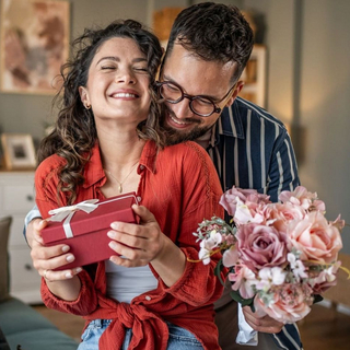Junger Mann &uuml;berrascht seine Freundin mit einem Geschenk und Blumen zu Hause