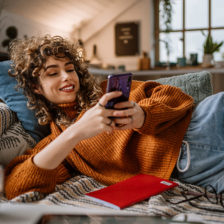 Junge Frau liegt auf ihrer Couch, w&auml;hrend sie ihr mobiles Smartphone benutzt
