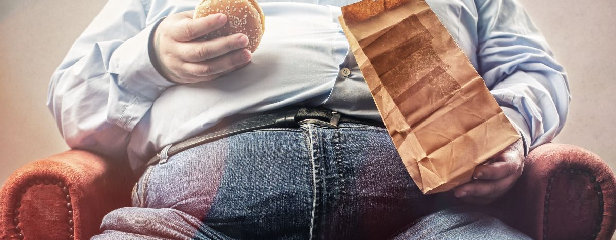 Dicker Mann sitzt auf dem Sofa und h&auml;lt einen Burger in der Hand.