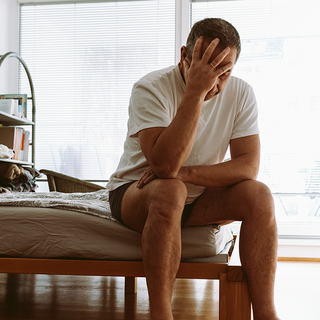 Mann sitzt an der Bettkante und st&uuml;tzt seinen Kopf mit der Hand ab