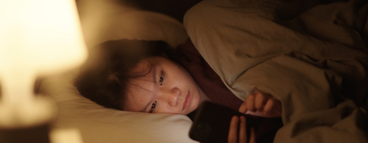 Ein asiatisches M&auml;dchen liegt im Bett und schaut auf ihr Smartphone.