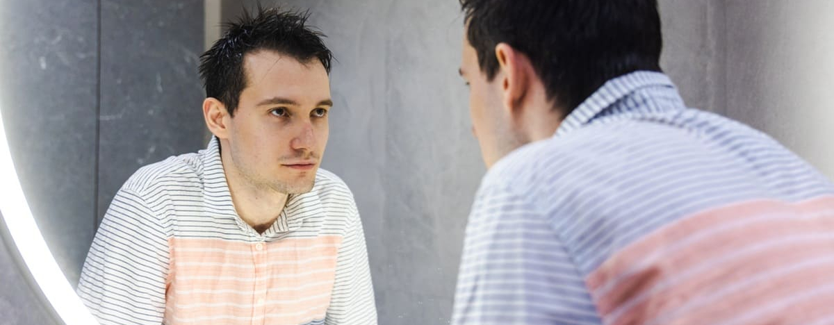 Ein junger Mann mit nassen Haaren beugt sich zu einem runden, beleuchteten Spiegel und betrachtet nachdenklich sein Spiegelbild in einem modernen Badezimmer.
