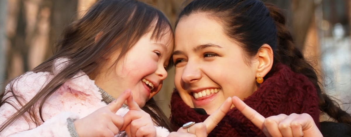 Ein M&auml;dchen mit Down-Syndrom und eine junge Frau spielen drau&szlig;en, l&auml;cheln sich an.