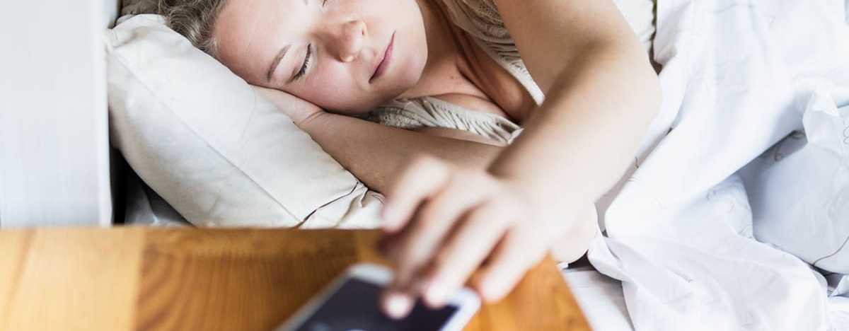 Junge Osteurop&auml;ische Frau liegt im Bett und schaltet morgens den Wecker auf ihrem Handy aus