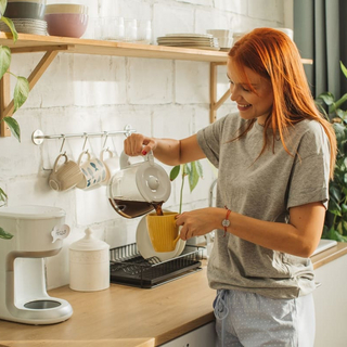 Eine junge Frau f&uuml;llt Kaffee aus einer Filtermaschine in eine Tasse.