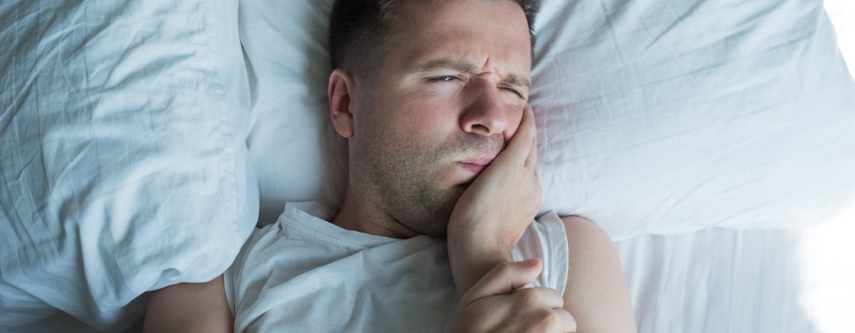 Mann liegt mit Zahnschmerzen im Bett und h&auml;lt sich eine Hand an die Wange