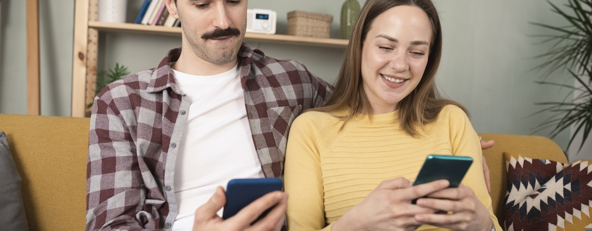 Junges Paar sitzt auf Sofa, beide haben ihr Smartphone in der Hand und chatten &uuml;ber den TI-Messenger mit einem Arzt.