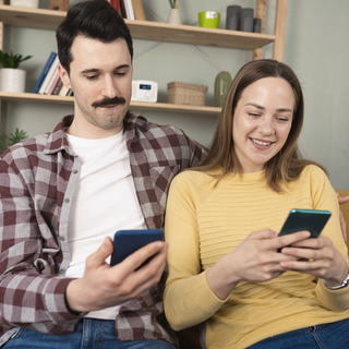 Junges Paar sitzt auf Sofa, beide haben ihr Smartphone in der Hand und chatten &uuml;ber den TI-Messenger mit einem Arzt.