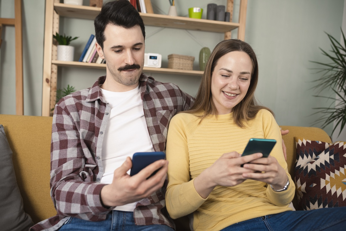 Junges Paar sitzt auf Sofa, beide haben ihr Smartphone in der Hand und chatten &uuml;ber den TI-Messenger mit einem Arzt.