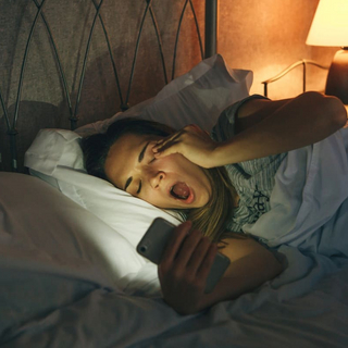 Ein M&auml;dchen liegt im Bett, starrt auf ihr Smartphone und g&auml;hnt
