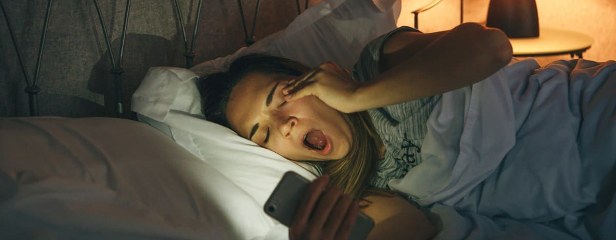 Ein M&auml;dchen liegt im Bett, starrt auf ihr Smartphone und g&auml;hnt
