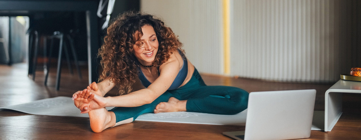 Eine junge Frau mit lockigen Haaren trainiert in ihrem Wohnzimmer auf einer Yogamatte vor ihrem Laptop.