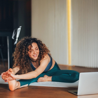 Eine junge Frau mit lockigen Haaren trainiert in ihrem Wohnzimmer auf einer Yogamatte vor ihrem Laptop.