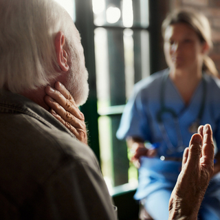 Ein &auml;lterer Herr, der mit einer &Auml;rztin spricht und sich dabei den Hals h&auml;lt.