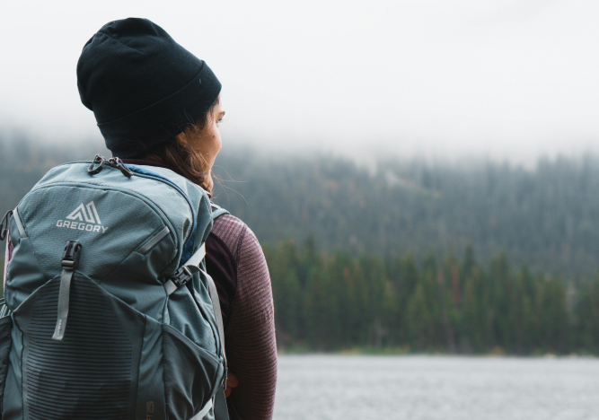 Eine junge Frau mit Rucksack und M&uuml;tze blickt auf einen See.