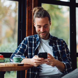 Mann mit Zopf sitzt entspannt im Caf&eacute; und freut sich dar&uuml;ber, was er auf seinem Smartphone sieht.