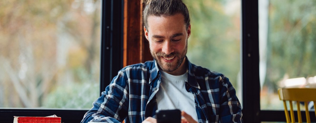 Mann mit Zopf sitzt entspannt im Caf&eacute; und freut sich dar&uuml;ber, was er auf seinem Smartphone sieht.