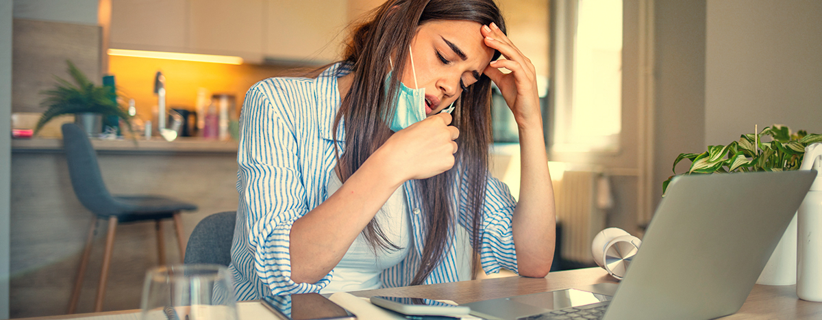 Eine Frau sitzt im Homeoffice, sie zieht sich die Maske vom Mund und h&auml;lt sich den Kopf vor Schmerzen