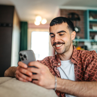 Ein Hipster-Mann sitzt auf einem Sofa in seinem Wohnzimmer und &auml;ndert seine pers&ouml;nlichen Daten am Smartphone