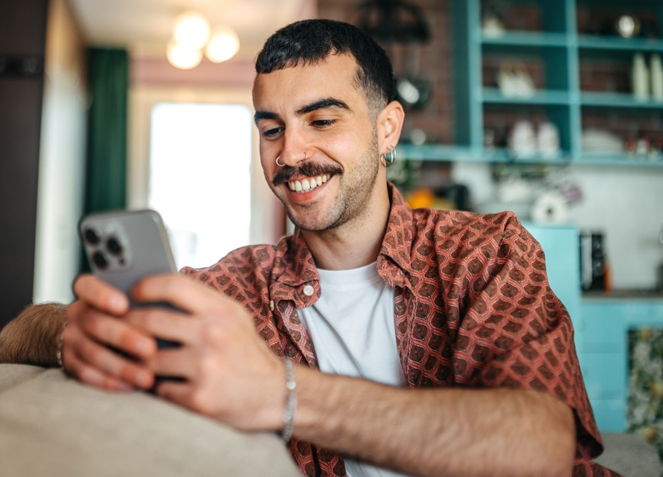 Ein Hipster-Mann sitzt auf einem Sofa in seinem Wohnzimmer und &auml;ndert seine pers&ouml;nlichen Daten am Smartphone