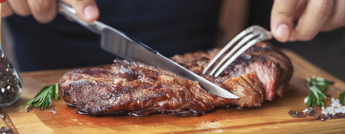 Ein St&uuml;ck Fleisch liegt auf einem Brett und wird mit Messer und Gabel geschnitten.