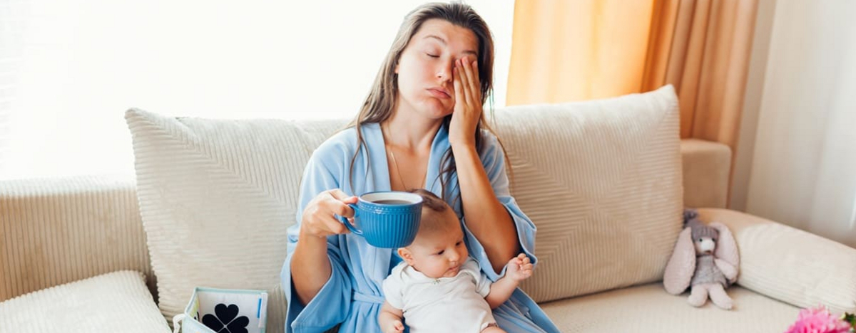 M&uuml;de, ersch&ouml;pfte junge Mutter, die Morgenkaffee trinkt, mit Neugeborenem, das zu Hause auf ihrem Scho&szlig; sitzt. Postpartale Depression