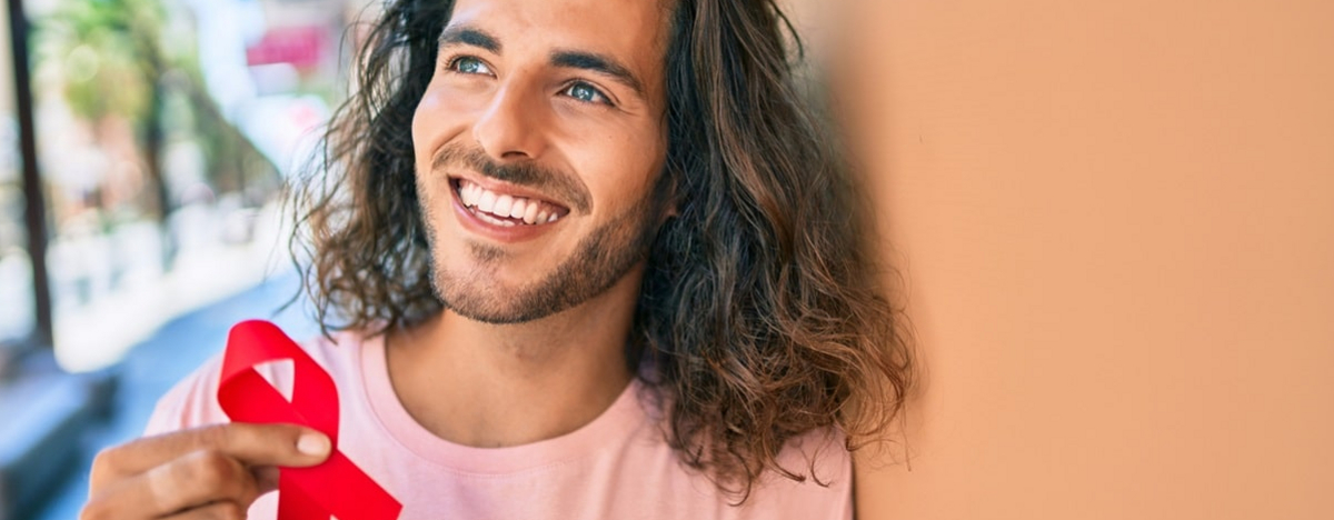 Ein junger Mann lehnt an einer Wand und h&auml;lt eine rote AIDS-Schleife in die Kamera.