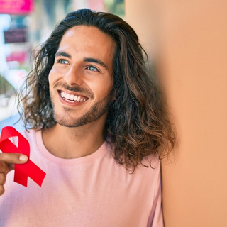 Ein junger Mann lehnt an einer Wand und h&auml;lt eine rote AIDS-Schleife in die Kamera.
